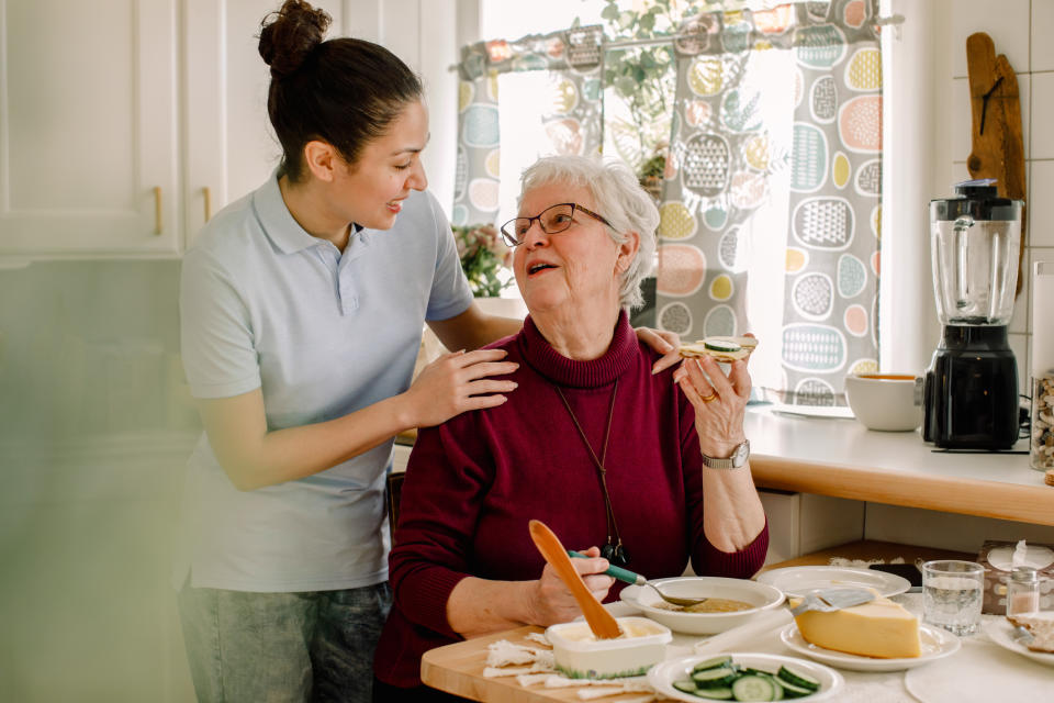 Young woman assists smiling elderly woman in the kitchen, with vegetables and cooking utensils on the counter