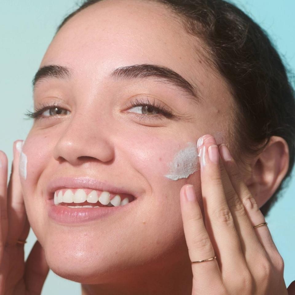 Person smiling, applying cream on cheek, promoting skincare product