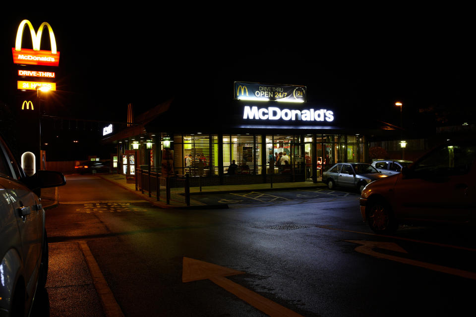 A McDonald's restaurant at night with a lit drive-thru sign and several cars in the parking lot. People are visible inside the restaurant