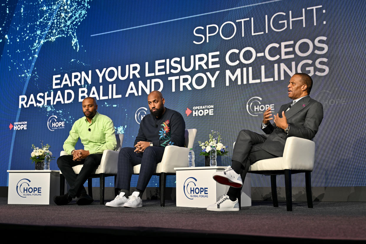 Rashad Bilal, Troy Millings and John Hope Bryant speak onstage with a banner in the background saying: Spotlight, Earn Your Leisure, Rashad Balil [sic] and Troy Millings, Operation Hope.