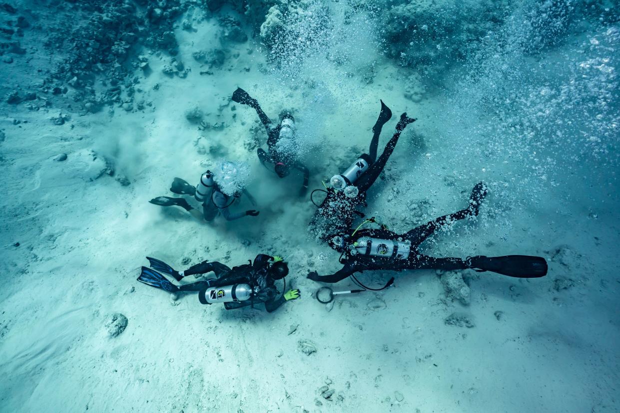 Five scuba divers in a circle on the seafloor looking at objects