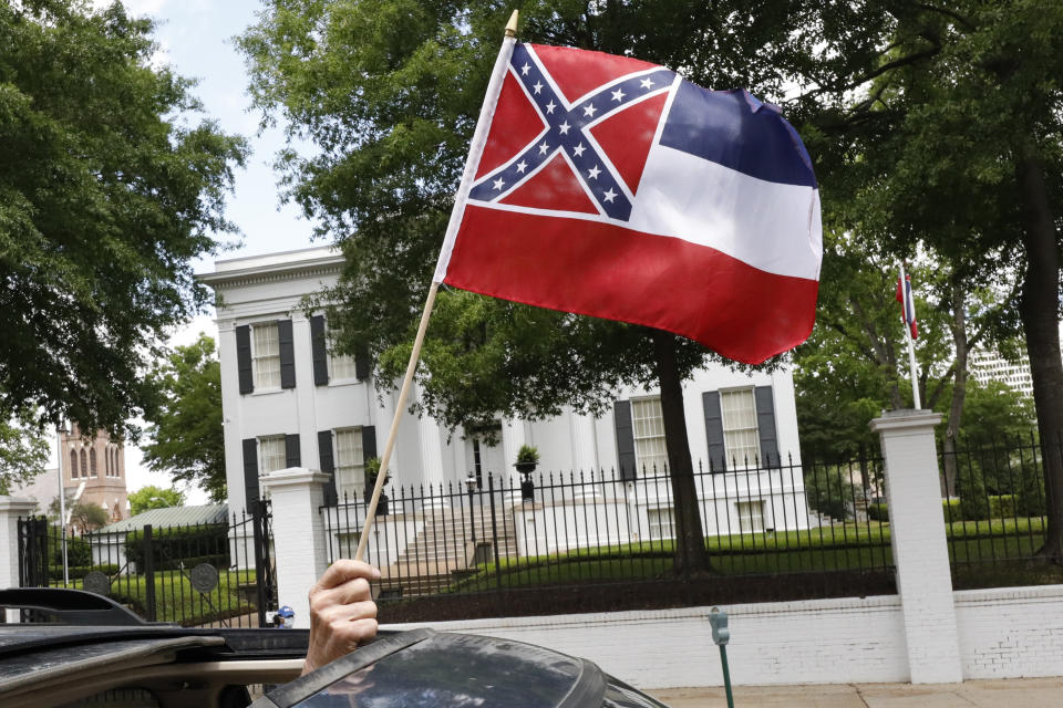 In this April 25, 2020 photograph, a small Mississippi state flag is held by a participant during a drive-by "re-open Mississippi" protest past the Governor's Mansion, in the background, in Jackson, Miss. This current flag has in the canton portion of the banner the design of the Civil War-era Confederate battle flag, that has been the center of a long-simmering debate about its removal or replacement. (AP Photo/Rogelio V. Solis)