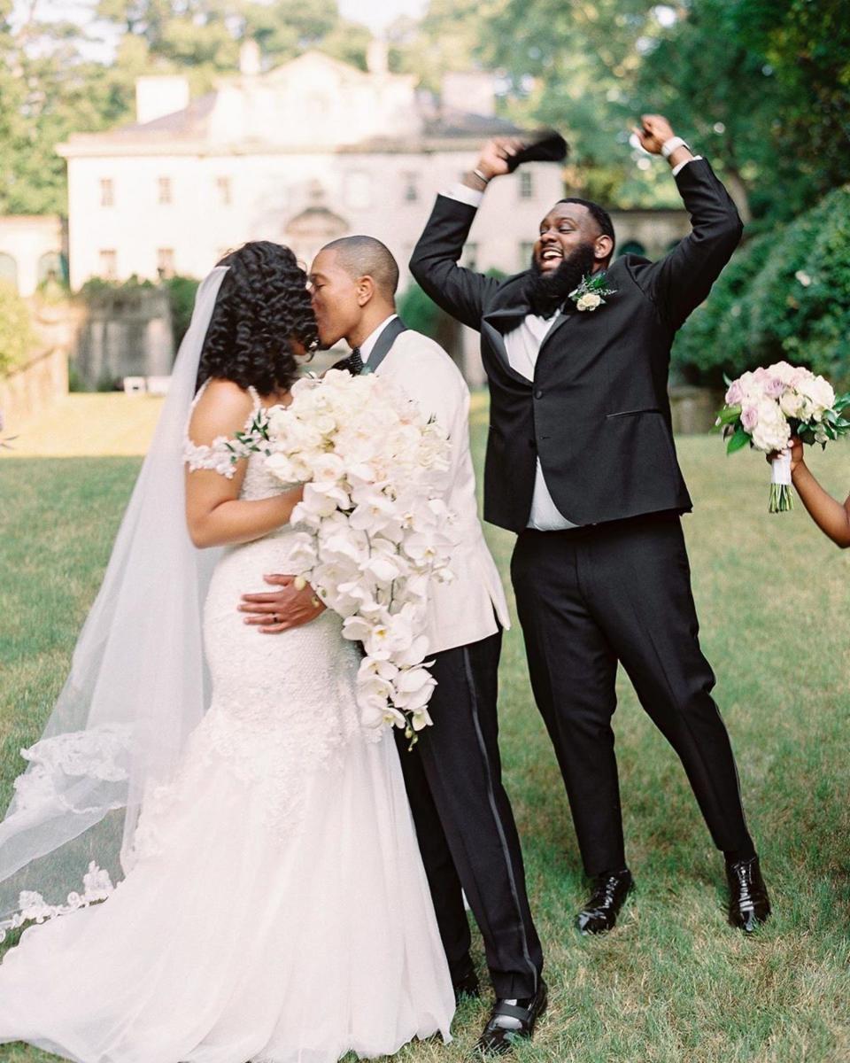 A bride and groom kiss as a groomsman jumps and cheers next to them.