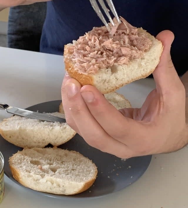 A person using a fork to spread tuna on a bread roll