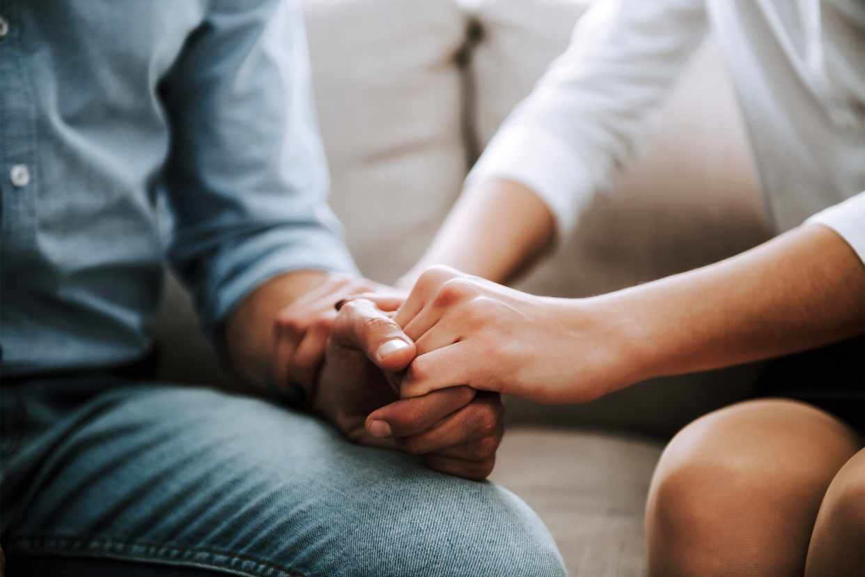 Focus on a couple's hands held together on the man's leg wearing jeans, midsection of both on a beige couch, man on the right, woman on the left