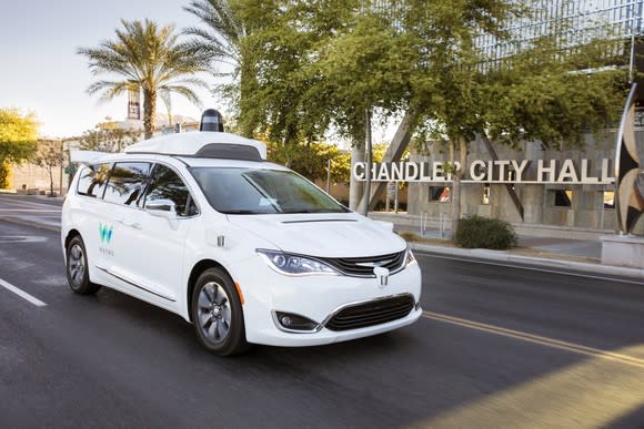 A white Chrysler Pacifica minivan with Waymo markings and visible self-driving sensor hardware is shown on a public road in Chandler, Arizona.