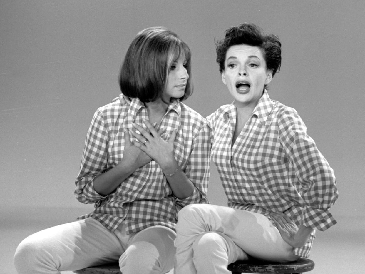In this black and white still from "The Judy Garland Show," Barbra Streisand on a stool next to Judy Garland, looking at her while Garland is either singing or speaking. Both are wearing gingham shirts, white pants, and white ballet flats.