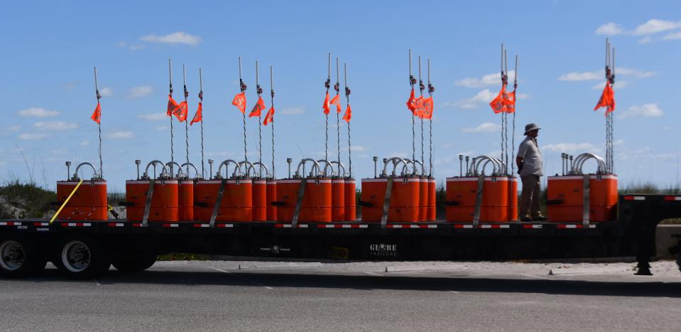 A truckload of mystery aquatic items are sitting on a tractor trailer bed in the parking lot of Patrick Space Force Base. The bright orange objects are numbered, have antenna and each one has two boat propellers protruding from them. 