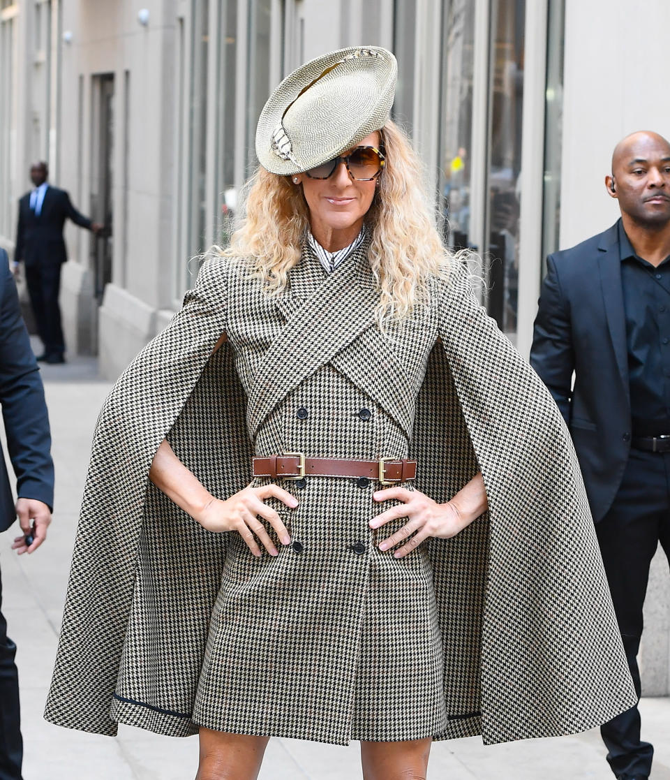 Celine Dion poses in a fashionable checkered cape dress with a matching hat and sunglasses, standing confidently with hands on hips