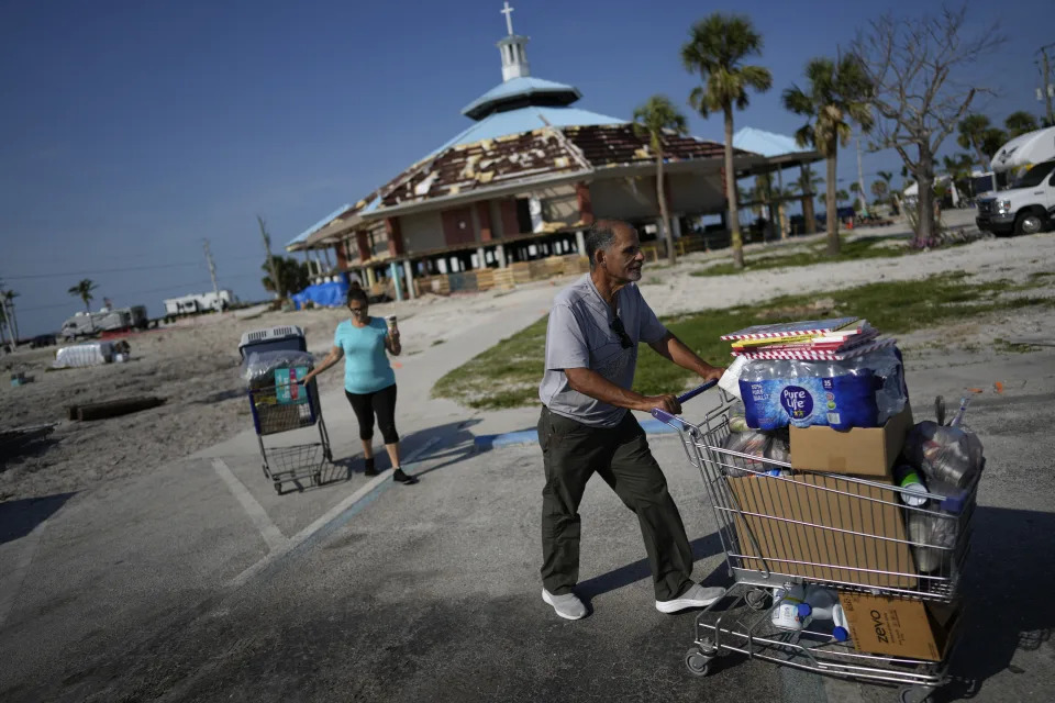 Omar Del Rio, a civil engineer currently subcontracted to FEMA, and his wife Maria wheel shopping carts full of groceries and supplies to their car as they leave the free food pantry operating underneath the heavily damaged Beach Baptist Church in Fort Myers Beach, Fla., Thursday, May 11, 2023. Before Hurricane Ian devastated Fort Myers Beach in 2022, the Del Rios rented an apartment on the island, living near the rented homes of their adult son and daughter, who each lived with their spouse and three children. All three homes were lost in the storm, and the six adults and six children were forced to spend months living together in one camper. (AP Photo/Rebecca Blackwell)