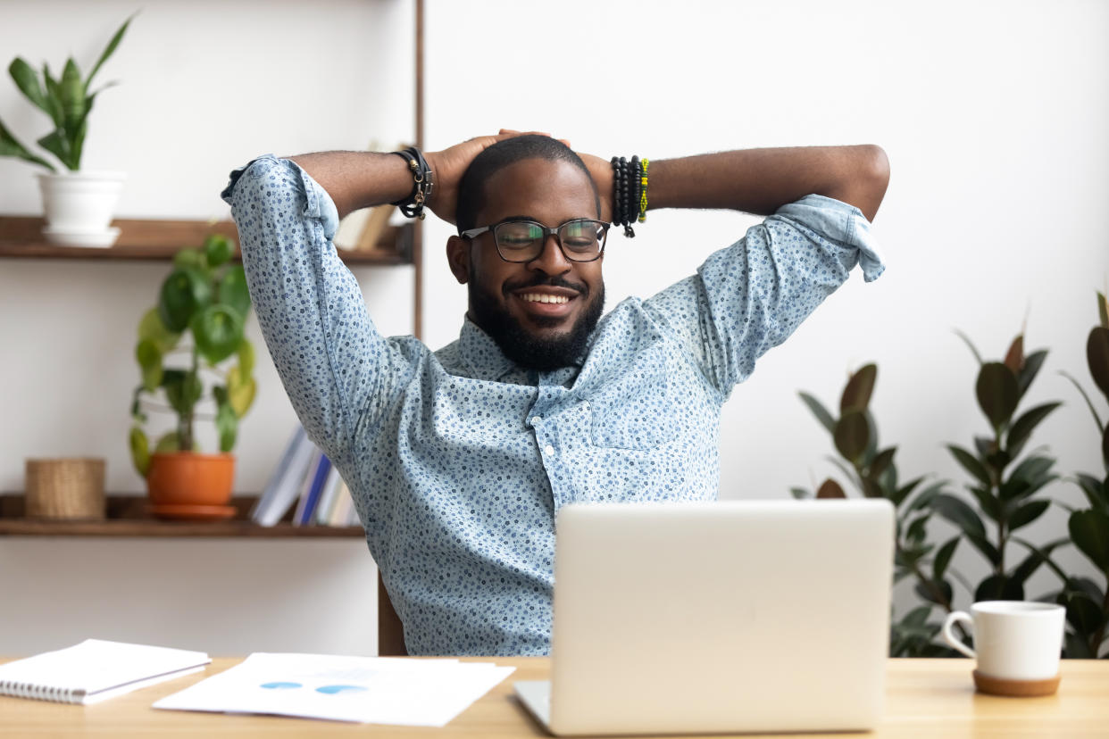 Smiling Afro-American businessman holding hands behind head sitting at office desk behind laptop. Happy black employee feeling no stress, relaxing, watching funny video after successful working
