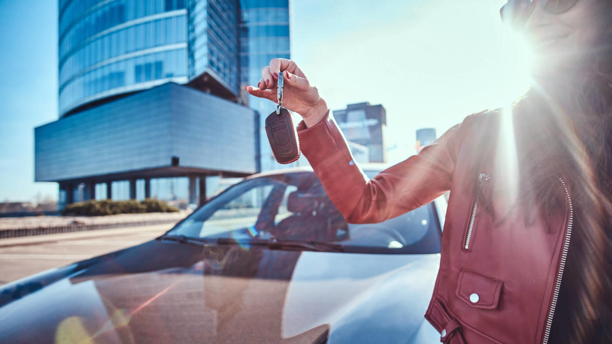 Happy young woman just got keys from her new car, which is behide.