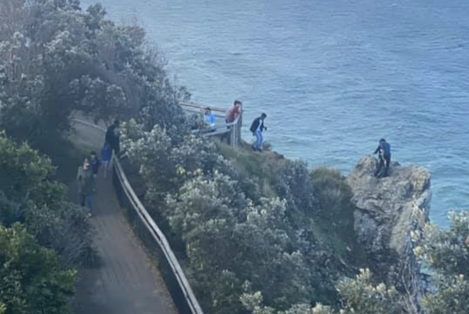 Pictured is a person standing on a cliff near Cape Byron Lighthouse while two others close by watch on. 