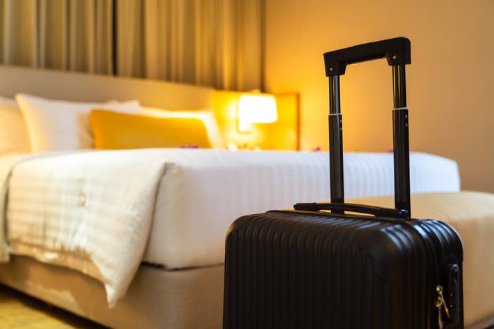 A suitcase with a telescopic handle stands in front of a neatly made hotel bed with white linens, a yellow pillow, and a bedside lamp