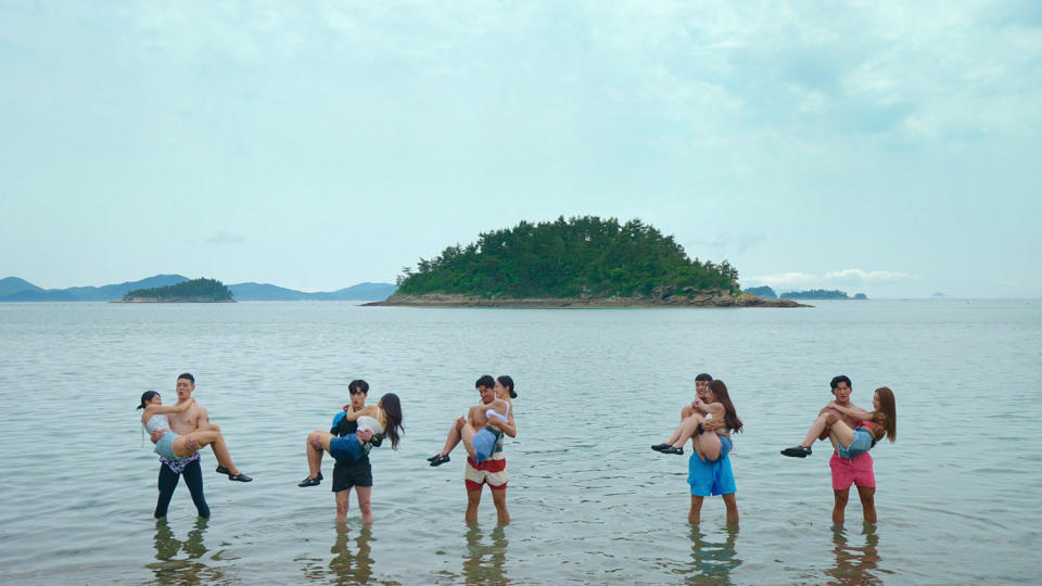 five men, each holding a woman in their arms, stand in the water in front of an island