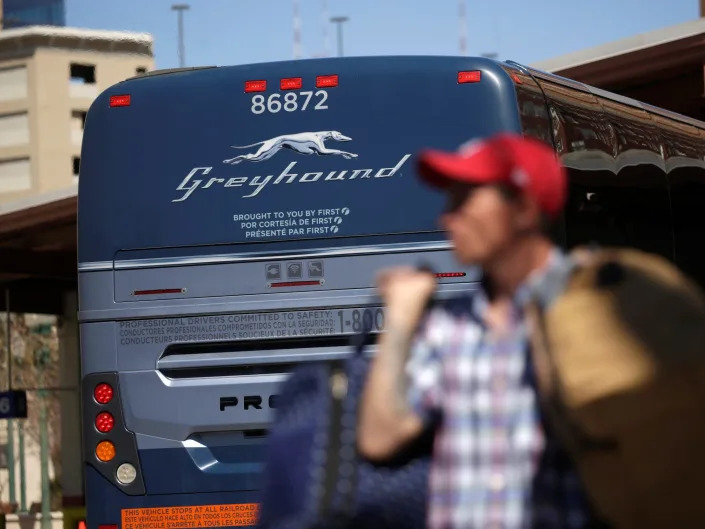 A person wearing a hat carrying a bag while a Greyhound bus is in the back.