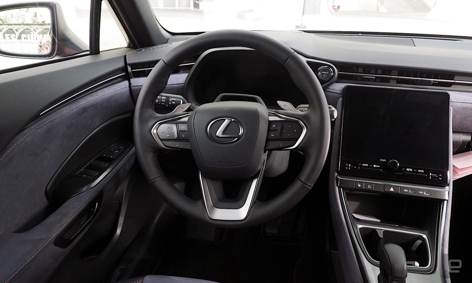 Bild der Fahrerposition in einem Lexus LBX mit dunkelgrauem und schwarzem Innenraum. Das Lenkrad steht vor einem digitalen Instrumentenkasten, rechts davon befindet sich ein Infotainmentsystem.