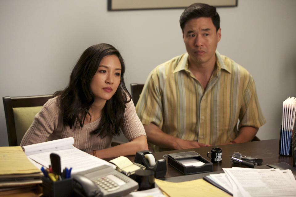 Constance Wu and Randall Park are sitting at a desk covered with papers, looking serious and focused