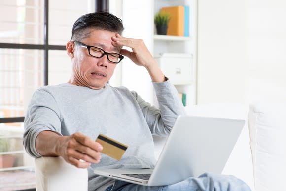 A worried man holding his credit card and looking at his laptop.