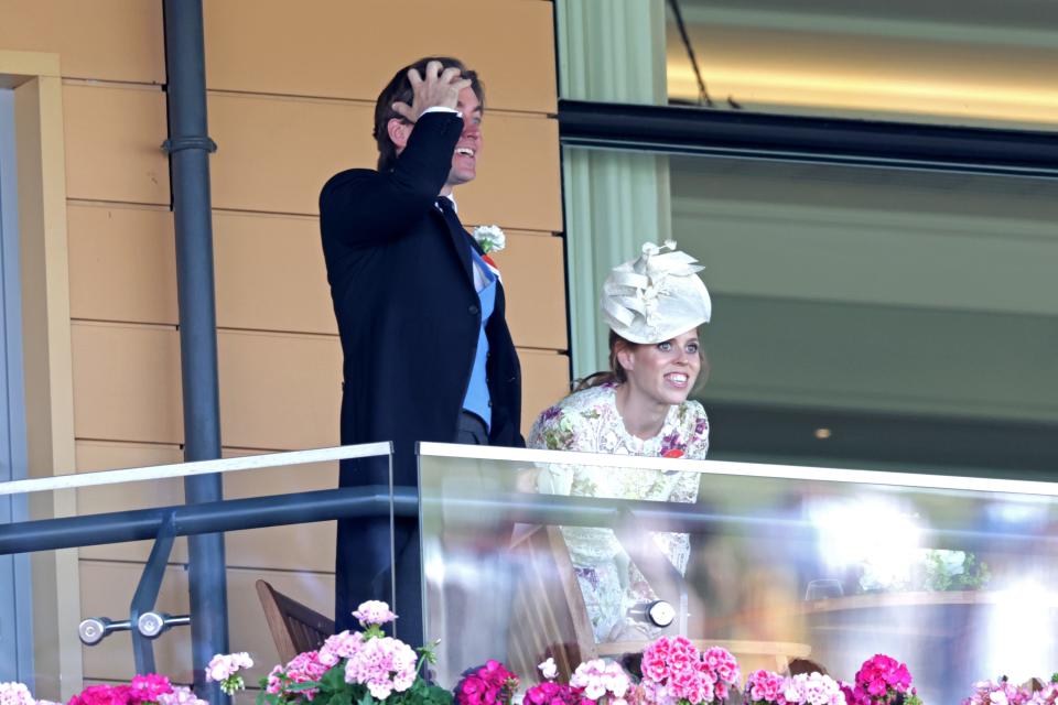 Edoardo Mapelli Mozzi and Princess Beatrice standing on a glass balcony that is lined with pink flowers while exclaiming.
