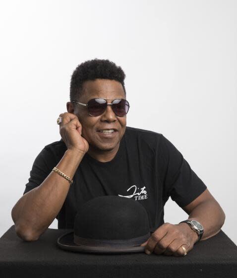 Tito Jackson in a black t-shirt and aviator sunglasses poses with his elbow on a table