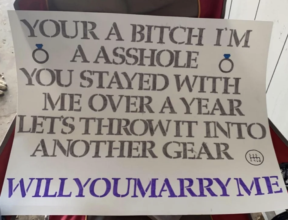 A sign says "you're a bitch, I'm an asshole, you stayed with me over a year, let's throw it into another gear. Will you marry me?"