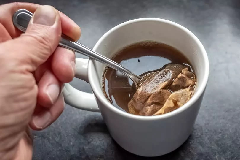 Human hand squeezing a teabag against the side of a mug with a silver teaspoon whilst making a mug of English tea