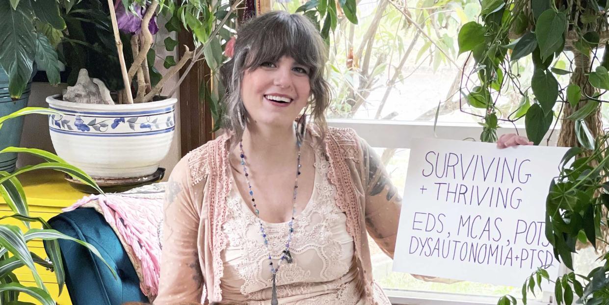 aunia kahn sitting in a white dress petting her dog, she carries a sign that says surviving and thriving with her invisible disabilities