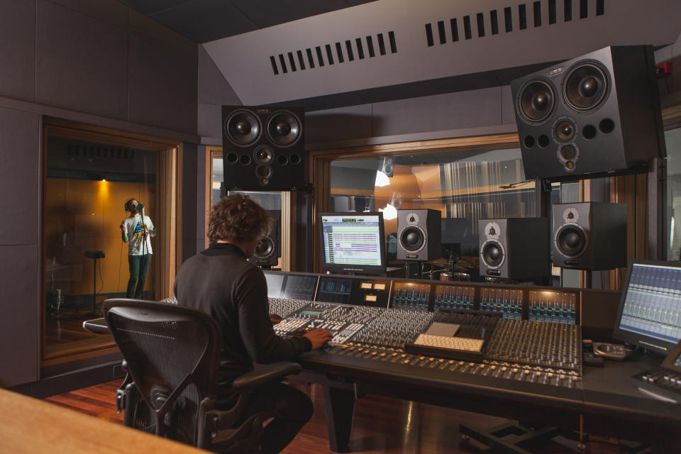 Person at mixing console in studio while another stands at mic in booth