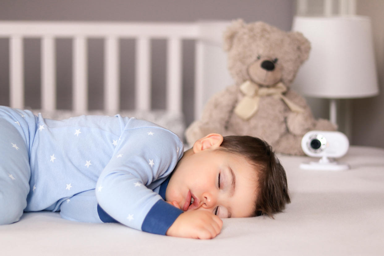 Cute little baby boy in light blue pajamas sleeping peacefully on bed at home with baby monitor camera and soft teddy bear toy at background. Child daytime sleeping schedule