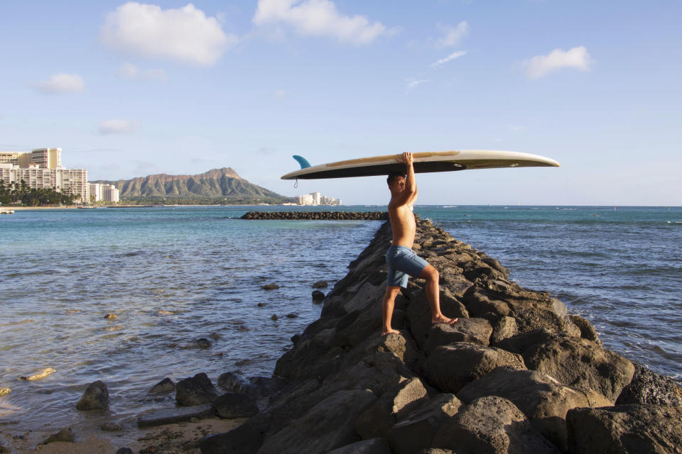 In this photo provided by Yoko Liriano, Bryant de Venecia poses for a photo with his paddleboard in Honolulu, Wednesday, Nov. 11, 2020. He started stand-up paddle-boarding when there were fewer tourists coming to Hawaii during the pandemic. He's among the Hawaii residents feeling ambivalence toward tourists returning now that the state is allowing incoming travelers to to bypass a 14-day quarantine with a negative COVID-19 test. (Yoko Liriano via AP)