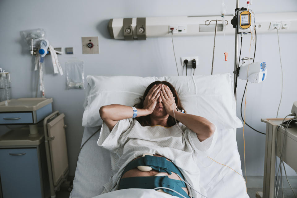 A woman in a hospital bed, wearing a hospital gown and medical equipment, covers her face with both hands. Medical devices are attached to her body and around the bed