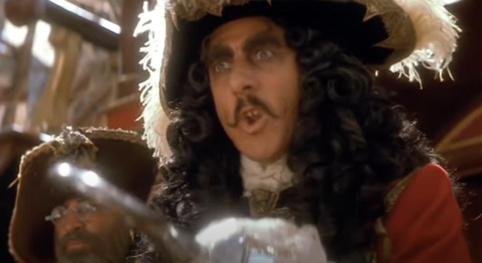 actor Dustin Hoffman wears a long black wig and a mustache as he holds a hook out in front of him