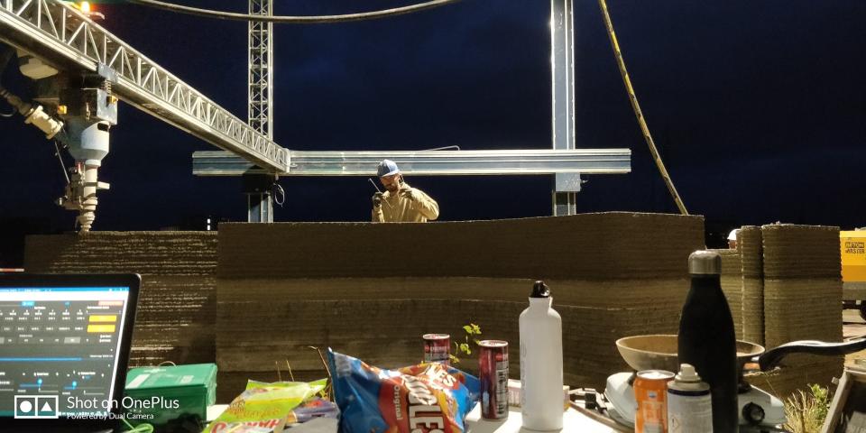 A 3D printer as it prints the walls of a tiny home at night. There's snacks and beverages in the foreground and somebody standing above the alls in the background.