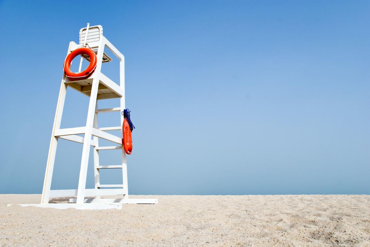 Empty white lifeguard chair on an empty beach with  orange life buoys hanging on the side.The chair stands against a clear blue sky in the sand.