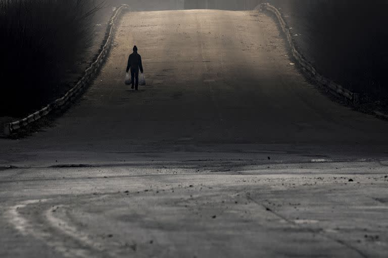 Un hombre lleva las compras mientras el humo de un almacén destruido por los bombardeos rusos proyecta sombras en la carretera a las afueras de Kyiv, Ucrania, el jueves 24 de marzo de 2022.