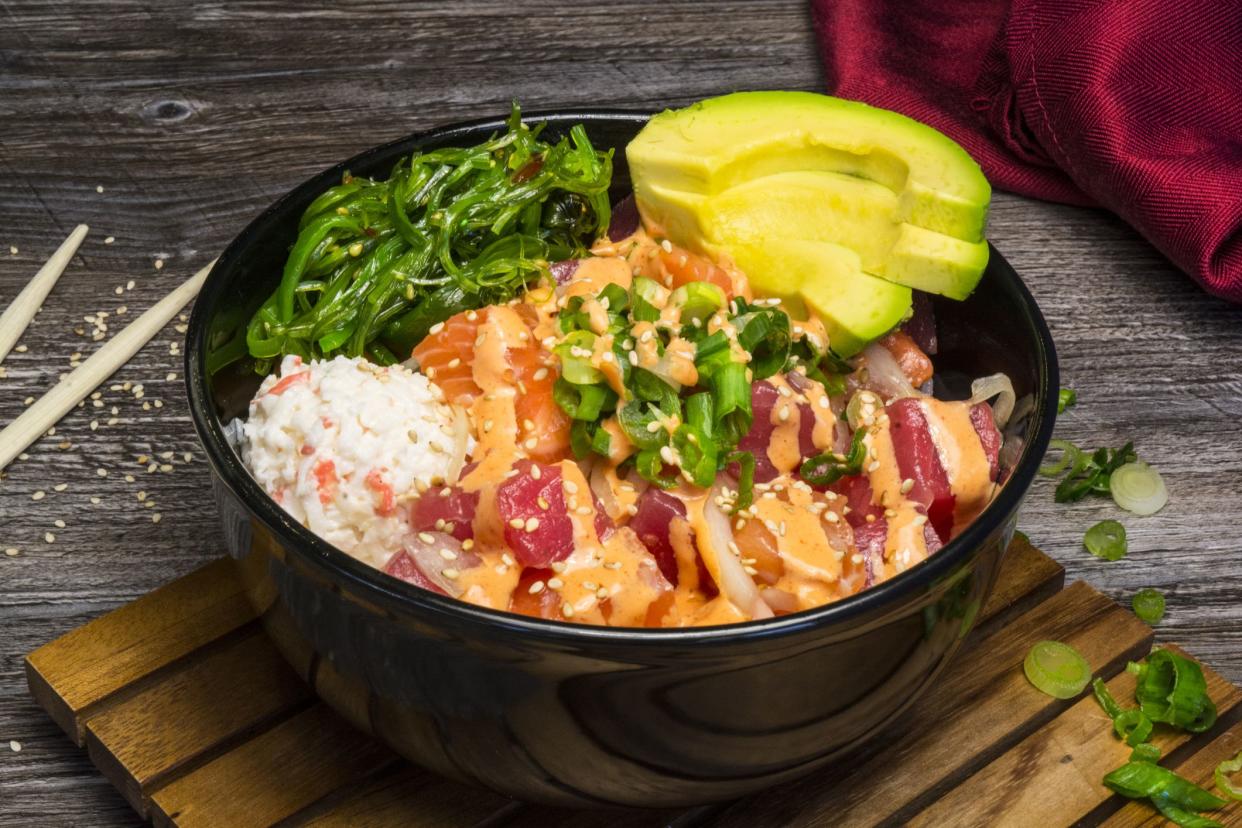 poke bowl with raw fish, avocado, sesame seeds, and algae in wooden surface with chopsticks and napkin in background