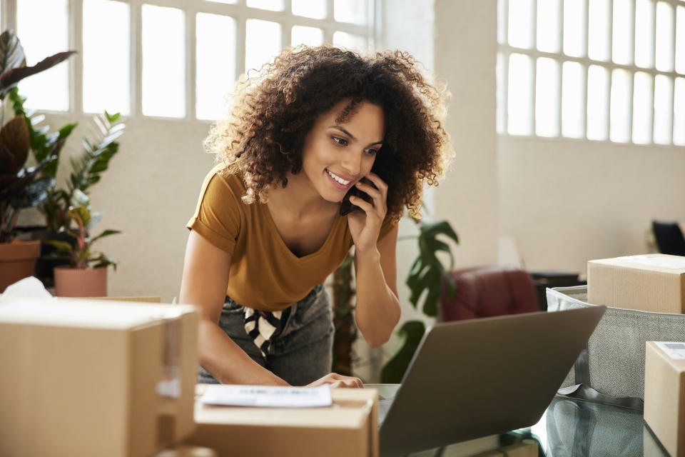 Young female talking on smart phone while using laptop. Confident businesswoman is working at home office. She is having curly hair.