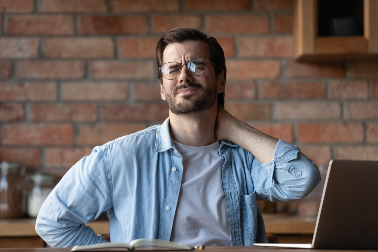 Tired man in glasses massaging rubbing stiff neck muscles, feeling pain, sitting at table, exhausted businessman freelancer suffering from discomfort after sedentary work, overworking concept