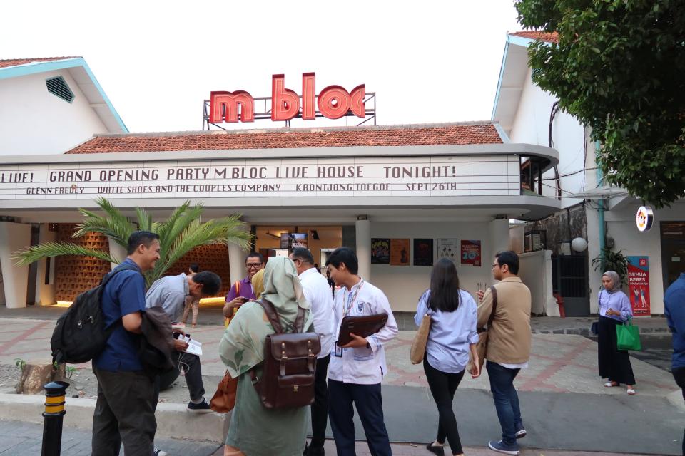 Crowds gather outside a white building with red letters saying "M Bloc"