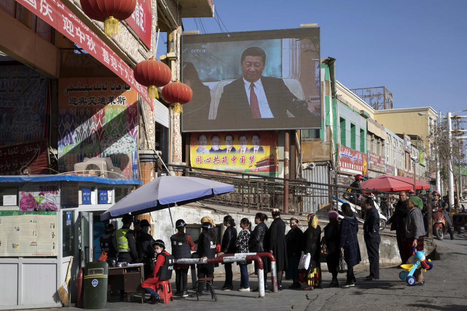 FILE - Residents walk through a security checkpoint into the Hotan Bazaar where a screen shows Chinese President Xi Jinping in Hotan in western China's Xinjiang region on Nov. 3, 2017. Within China, the Communist Party under Xi has increased surveillance, tightened already strict control over speech and media and cracked down further on dissent, censoring even mildly critical views and jailing those it believes went too far. Authorities have repressed predominantly Muslim groups in China's Xinjiang region in a harsh anti-extremism campaign that has raised a human rights outcry internationally. (AP Photo/Ng Han Guan, File)