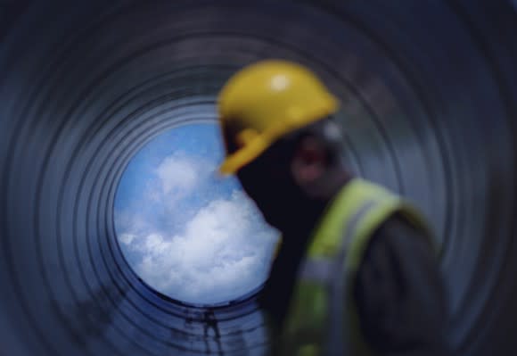 A pipeline construction worker walking past a pipe with a blue sky in view.