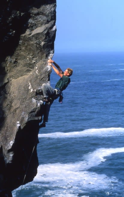 Man climbs steep prow of grey rock by a back drop of ocean.