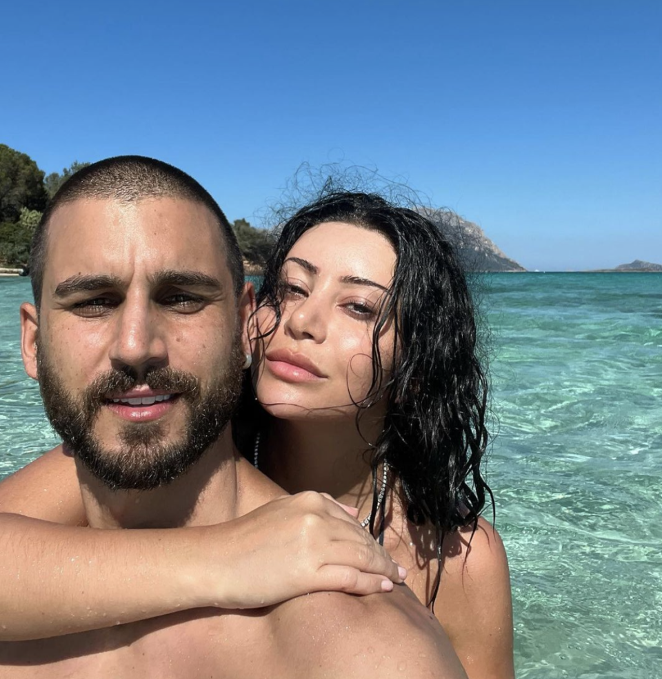 Martha and Michael Brunelli pictured in clear blue water with rocks behind. She stands behind him with her arm around his neck while he takes a selfie.