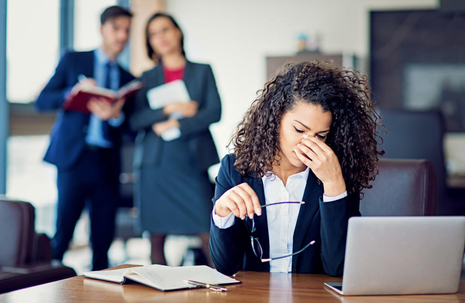 A sad woman sitting with her hand on her face while two coworkers sneer at her from behind