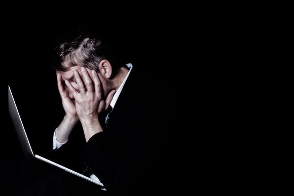 Dark-suited businessman in a dark room, leaning on his laptop with his head in his hands.