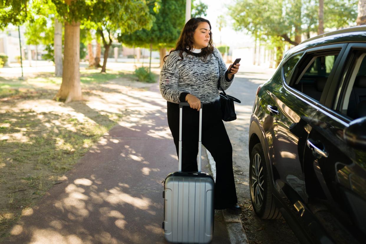 Fat latin woman with a suitcase waiting for her taxi car while requesting a trip on a ride share app