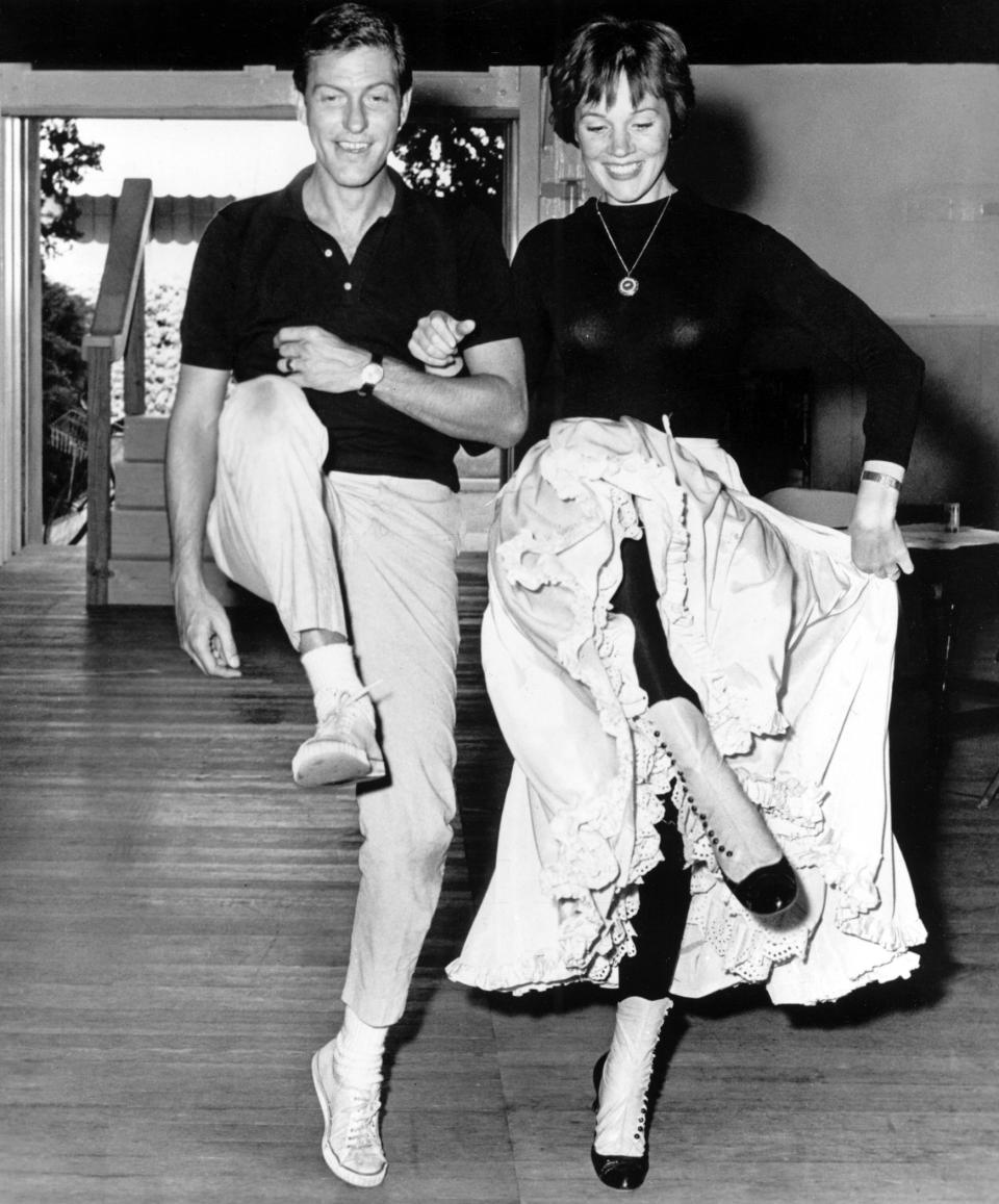 Dick Van Dyke and Julie Andrews dance together on a wooden floor, both mid-step with broad smiles. Julie's skirt is lifted to reveal her footwear