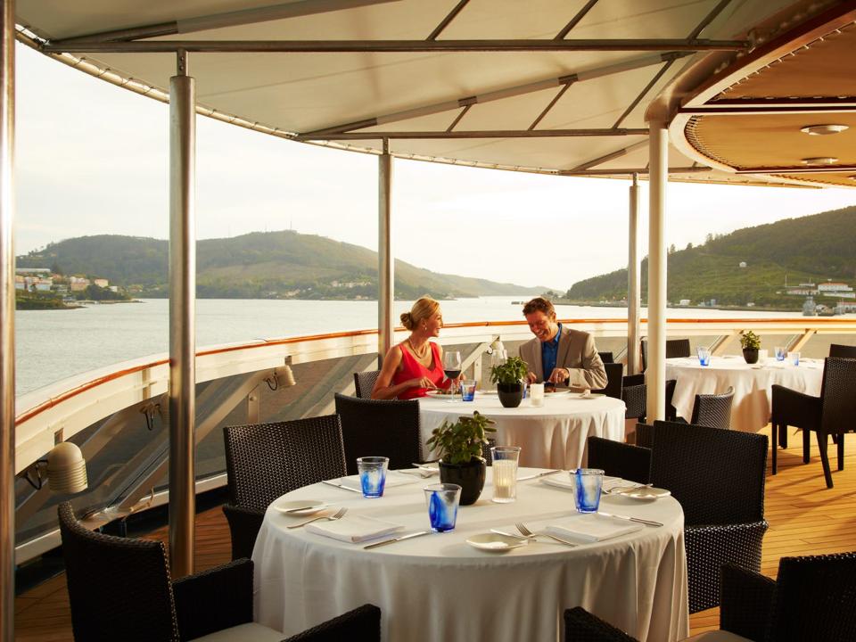 A couple sitting at a dining table on a deck that overlooks a coastline.