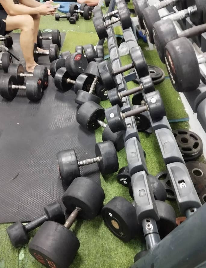 Dumbbells scattered across a gym floor next to a rack, with a person's legs visible, suggesting disorganization